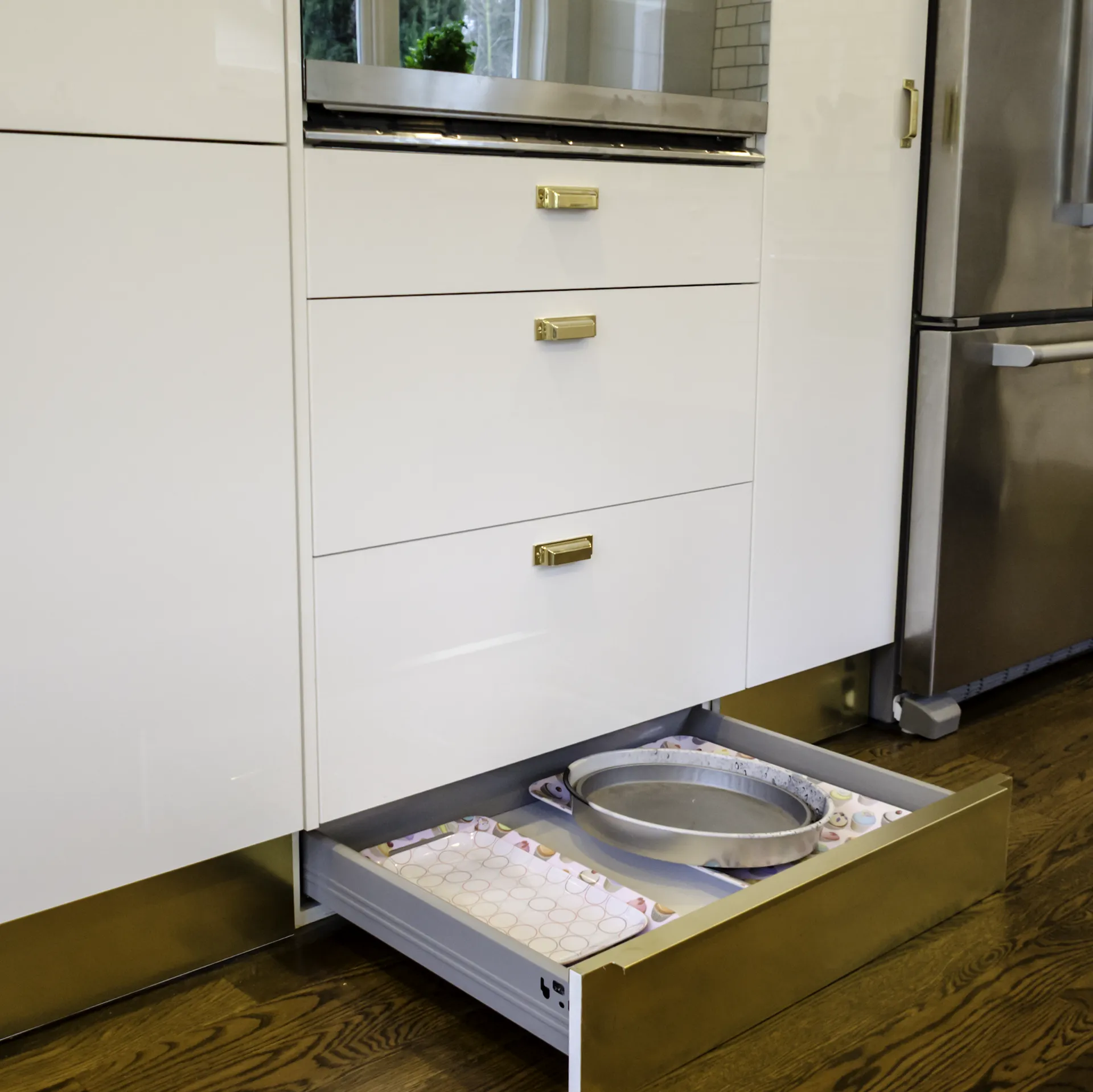 IKEA toe-kick drawer pulled open showing bakeware storage under white gloss cabinets with gold hardware